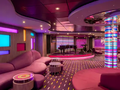 Moderne Purple Jazz Bar an Bord der MSC Splendida. Lila Sofas, Bühne mit Klavier, farbige Leuchtflächen an Wänden und Säulen. Gemusterter Teppich.