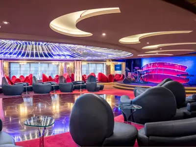 Elegante Aft Lounge an Bord der MSC Splendida. Eine Bühne mit Musikinstrumenten, umgeben von roten und schwarzen Loungesesseln, prägt den Raum. Glänzende Böden und kunstvolle Deckenbeleuchtung erzeugen eine festliche Stimmung.
