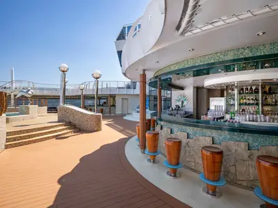 Blick auf die Bar del Riccio an Bord der MSC Splendida bei Tageslicht. Die geschwungene Outdoor-Bar mit hellblauen Mosaiken und holzfarbenen Barhockern steht auf dem oberen Deck des Kreuzfahrtschiffs, umgeben von weiterer Deckgestaltung und einer Skulptur.