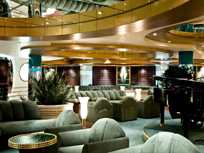 Die La Prua Piano Bar an Bord der MSC Splendida. Grüne Samtsofas, goldene Tische und ein schwarzer Flügel. Elegante Beleuchtung und Pflanzen prägen das luxuriöse Ambiente.