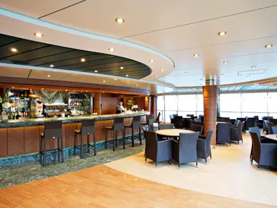 Moderner Bar- und Loungebereich der MSC Splendida mit grünem Marmor, Holzwänden und einem Barkeeper. Bequeme Korbstühle an Tischen und der Theke vor heller Fensterfront.