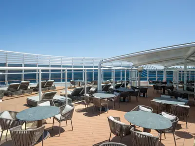 Das sonnige Außendeck der MSC Splendida zeigt moderne Tische, Stühle und Sonnenliegen unter einem Sonnensegel. Im Hintergrund sind das blaue Meer und eine Stadtsilhouette sichtbar. Keine Personen zu sehen.