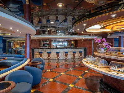 Elegante Top Sail Lounge an Bord der MSC Splendida. Sichtbar sind eine lange Bar, blaue Sitzgruppen und ein Buffet mit Gebäck und Snacks. Luxuriöses Ambiente mit spiegelnder Decke und Holzboden.