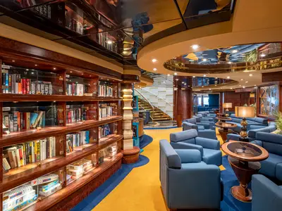 Blick in die elegante Bibliothek an Bord der MSC Splendida. Helle, glaenzende Holzregale mit Buechern und glaeserner Decke. Blaue Sitzgruppen mit Tischen, warme Beleuchtung und eine Treppe im Hintergrund.