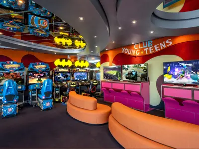 Blick in den lebhaften Teens Club der MSC Splendida mit orangefarbenen Sitzgelegenheiten, mehreren Bildschirmen für Videospiele und Arcade-Automaten wie 'Overdrive' und 'Batman'.