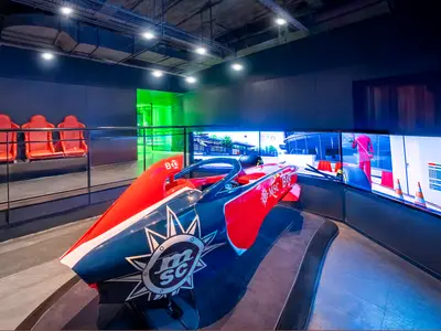 Ein roter und blauer MSC Formula Racer Rennsimulator steht an Bord der MSC Splendida vor drei großen Bildschirmen, die eine Rennstrecke zeigen. Daneben sind rote Rennsitze für Zuschauer.