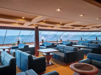 Eine stilvolle Lounge an Bord der MSC Splendida mit Reihen von blauen Sofas und Tischen. Große Fenster bieten einen Panoramablick auf eine belebte Küstenstadt und das Meer.