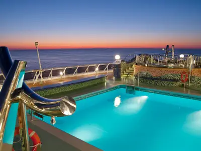 Eine malerische Abendansicht des beleuchteten Poolbereichs an Bord der MSC Splendida. Der türkisfarbene Pool wird von Unterwasserleuchten erhellt, umgeben von kunstvollen Mosaiken und einem Whirlpool im Hintergrund. Über dem ruhigen Meer erstreckt sich ein dramatischer, rot-oranger Sonnenuntergangshimmel.
