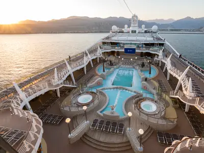 Der Aqua Park der MSC Splendida am Abend. Von oben sind Pools, Whirlpools und Liegestühle auf dem Deck zu sehen. Im Hintergrund das Meer, beleuchtet vom Sonnenuntergang über Bergen. Wenige Passagiere.