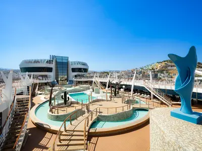 Das weitläufige Sonnendeck der MSC Splendida zeigt den Aqua Park mit Pools, Holzbrücken und Kaktusskulpturen. Eine blaue, abstrakte Statue steht rechts. Im Hintergrund ist eine bunte Küstenstadt zu sehen.