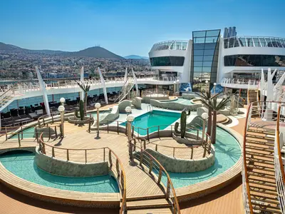 Der Aqua Park an Bord der MSC Splendida zeigt Pools, Brücken, Palmen und Kakteen. Im Hintergrund sind eine Küstenstadt und Berge unter blauem Himmel zu sehen.