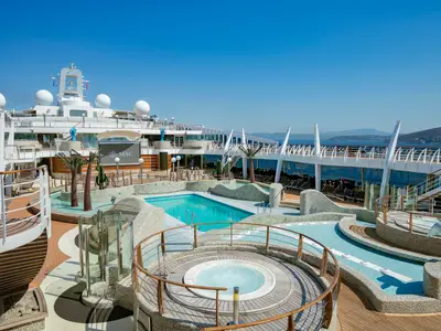 Blick auf den weitläufigen Aqua Park der MSC Splendida mit mehreren Pools, Whirlpool und künstlichen Palmen. Im Hintergrund blaues Meer und Berge. Keine Personen.
