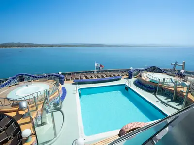 Eine helle Poollandschaft auf dem Sonnendeck der MSC Splendida unter blauem Himmel. Zu sehen sind ein großer Pool, zwei Whirlpools und Liegestühle, mit Blick auf das weite Meer und eine grüne Küste am Horizont. Eine panamaische Flagge weht im Wind.