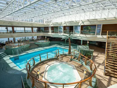 Innenpool L'Equatore der MSC Splendida: Großer Pool, Whirlpool, Palmenattrappe, Mosaikfliesen und Glasdach. Mehrere Lounge-Decks sind sichtbar.