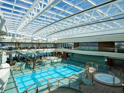 Weitwinkelansicht des hellen L'Equatore Indoor Pools an Bord der MSC Splendida. Ein grosses Glasdach, ein Hauptpool, Whirlpools und mehrere Decks mit Sitzbereichen sind zu sehen.