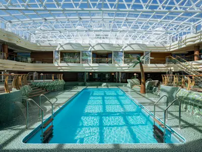 L'Equatore Indoor Pool der MSC Splendida: Heller Pool unter Glasdach. Türkisblaues Wasser, grüne Mosaikfliesen, Palmenattrappe, Whirlpools. Obere Decks mit Wandmosaiken.
