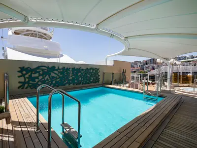 Ein sonniger Poolbereich an Bord der MSC Splendida. Der türkisfarbene Pool ist von einem Teakholzdeck umgeben, mit stilvoller Wanddekoration und Blick auf eine Küstenstadt.