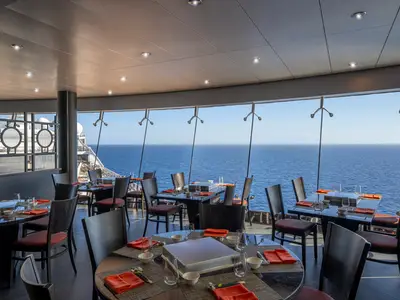 Modernes Spezialitätenrestaurant der MSC Splendida. Dunkle Tische, rote Akzente, grosse Fenster mit weitem Meerblick. Links ein Teil des Schiffdecks.