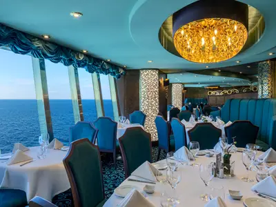 Blick in das elegante Villa Verde Restaurant an Bord der MSC Splendida. Gedeckte Tische und bequeme Sessel mit grossen Fenstern bieten einen weiten Blick auf das offene Meer unter blauem Himmel. Eine grosse goldene Leuchte schmückt die Decke.
