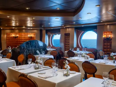 Innenansicht des eleganten Restaurants "La Reggia" an Bord der MSC Splendida. Zahlreiche Tische mit weissen Tischdecken und braunen Holzstühlen sind mit Geschirr und Gläsern gedeckt. Im Hintergrund sind drei grosse runde Fenster (Bullaugen) zu sehen, die einen weiten Blick auf das blaue Meer ermöglichen. Das Dekor ist klassisch mit hölzernen Wänden, Polsterungen und Wandleuchten.