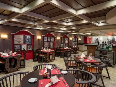 An Bord der MSC Splendida: Rustikales Spezialitätenrestaurant im Western-Stil. Viele Tische mit roten Platzsets, dunkle Holzstühle und eine Bar mit Spirituosen sind zu sehen.