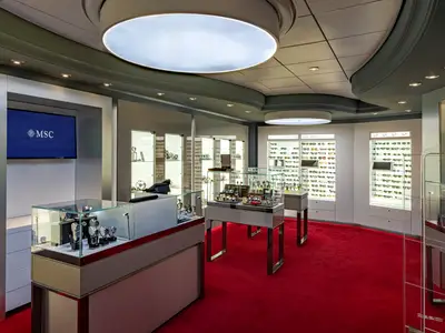 Ein heller, moderner Shop an Bord der MSC Splendida, ausgestattet mit gläsernen Vitrinen für Uhren und wandmontierten Displays für Sonnenbrillen. Der Boden ist mit einem leuchtend roten Teppich ausgelegt. Ein Bildschirm zeigt das MSC-Logo.