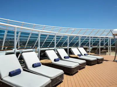 Sonnendeck auf der MSC Splendida: Elegante Lounge-Liegen mit blauen Handtüchern und Holztischen. Glaswände im Hintergrund zeigen eine Stadtsilhouette und Meer. Blauer Himmel.