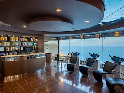 Eleganter Haarsalon des MSC Aurea Spa an Bord der MSC Splendida mit grosser Fensterfront zum Meer. Schwarze Stühle, Waschsessel und ein Produktregal sind zu sehen.