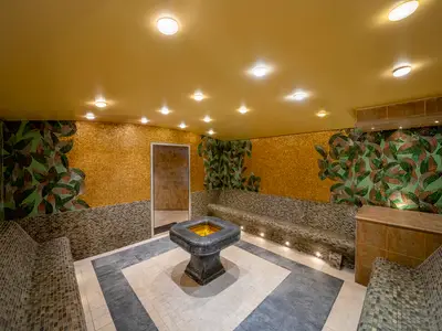 Ein Thermalbereich im MSC AUREA SPA an Bord der MSC Splendida. Der Raum ist mit kunstvollen Mosaiken verziert, die goldene Flächen und grüne Blätter zeigen. Integrierte Sitzbänke laden zur Entspannung ein. Ein zentrales Becken mit goldener Beleuchtung ist im Vordergrund zu sehen.