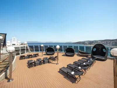 Das sonnige Sonnendeck der MSC Splendida mit Liegestühlen, runden Lounge-Sesseln und stilvollen Kugelleuchten auf dem Holzdeck. Im Hintergrund sind das weite Meer, eine bergige Küstenlandschaft und eine Stadt unter klarem, blauem Himmel sichtbar, ohne Personen.