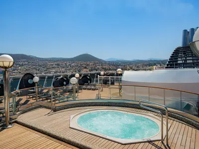 Auf dem Sonnendeck der MSC Splendida ist ein blauer Whirlpool zu sehen, umgeben von Holzplanken und Lounge-Möbeln, mit Blick auf eine bergige Küstenstadt.