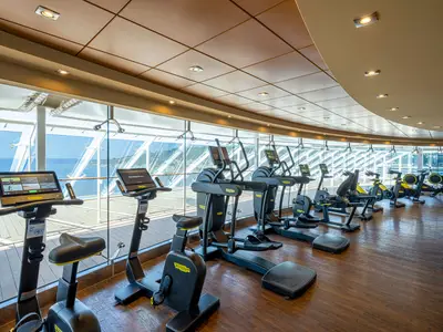 Ein moderner Fitnessraum an Bord der MSC Splendida. Die Ausstattung umfasst mehrere Technogym-Heimtrainer und Ellipsentrainer mit Bildschirmen. Große Panoramafenster bieten eine weite Aussicht auf das blaue Meer und eine grüne Küstenlandschaft unter klarem Himmel. Der Boden ist aus Holz und die Decke ist hellbraun mit integrierten Lichtern. Der Raum ist menschenleer und gut beleuchtet.