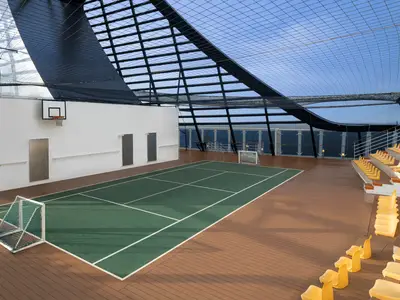 Blick auf die MSC Arena der MSC Splendida: Ein überdachter, grüner Sportplatz mit Basketballkorb und Fußballtoren. Gelbe Sitzreihen und Holzboden umgeben den Platz, mit Meerblick.