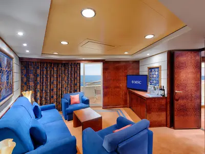 Eine luxuriöse Suite an Bord der MSC Splendida, ausgestattet mit blauen Sofas und Sesseln, einem Couchtisch und einem Fernseher. Ein Balkon mit weitem Meerblick ist sichtbar.