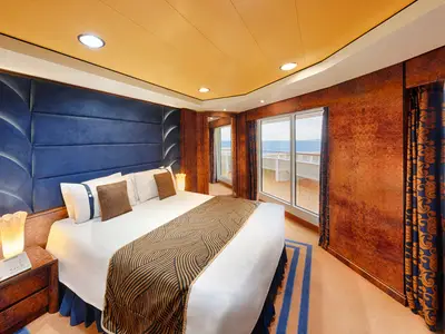 Eine geräumige Grand Suite Aurea an Bord der MSC Splendida. Ein großes Doppelbett mit blauer Kopflehne und einem gemusterten Läufer dominiert den Raum. Rechts öffnet sich eine Glastür zu einem privaten Balkon mit Meerblick. Dunkle Holzvertäfelungen und goldene Akzente prägen das elegante Interieur.