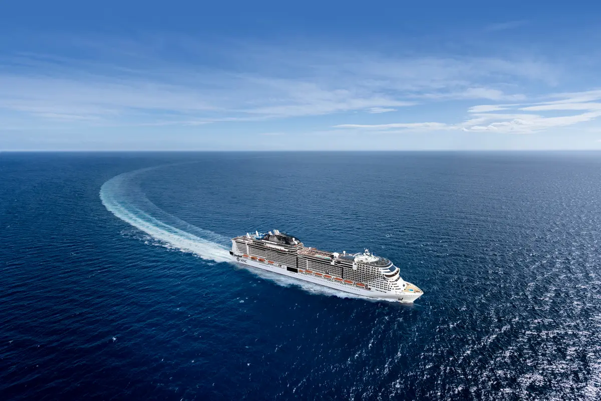 Vogelperspektive der MSC Virtuosa, die auf dem tiefblauen Meer eine weite Kurve fährt. Hinter dem Schiff ist eine lange, schäumende Heckwelle sichtbar, während der Himmel leicht bewölkt ist.