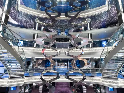Das Infinity Atrium an Bord der MSC Virtuosa. Glänzende, geschwungene Treppen und eine abstrakte Lichtskulptur in einem modernen Raum mit blauen Akzenten und sternenähnlicher Deckenbeleuchtung.
