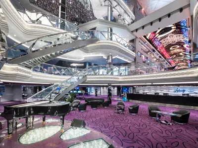 Das Infinity Atrium der MSC Virtuosa zeigt eine glänzende Grand Piano auf violettem Teppich, eine doppelte Wendeltreppe aus Glas und Edelstahl, moderne Sitzbereiche und eine beeindruckende digitale Decke mit künstlerischen Mustern.