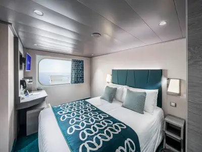Helle Aussenkabine der MSC Virtuosa mit Doppelbett, tealgrünem Kopfende und Musterbettdecke. Links ein Schreibtisch mit TV, rechts ein Fenster mit Meerblick. Modernes Design.