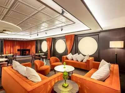 Blick auf die L'Atelier Bar & Art an Bord der MSC Virtuosa. Die Lounge ist mit orangen Sofas, Tischen mit hellgrünen Lampen und einer Bühne mit Klavier und roten Vorhängen ausgestattet.