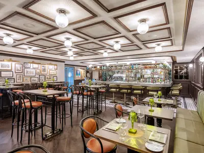 Elegante Bar an Bord der MSC Virtuosa mit hohen Tischen, braunen Lederstühlen und einer langen Theke mit Barhockern. Gerahmte Kunstwerke zieren die Wände.