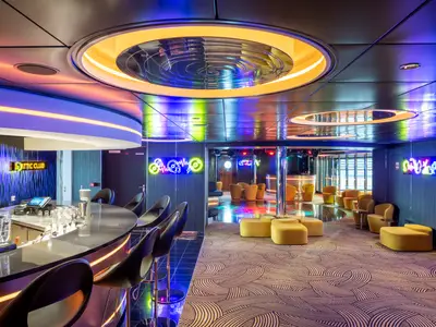 Der lebhafte Attic Club an Bord der MSC Virtuosa. Links eine Bar mit Hockern, rechts eine Tanzfläche mit leuchtenden Möbeln und Neon-Schildern an der Wand. Die Decke zeigt Spiegel und farbige Lichtinstallationen.