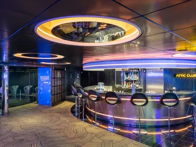 Der moderne Attic Club an Bord der MSC Virtuosa. Eine beleuchtete Bar mit Barhockern. Futuristische Deckenleuchten und Neonlichter schaffen eine lebhafte Atmosphäre.