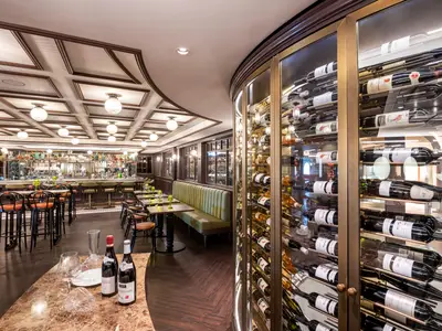 L'Atelier Bar an Bord der MSC Virtuosa mit einem runden Weinkeller rechts. Links eine lange Bar und Tische. Im Vordergrund Weinflaschen und Gläser auf einem Marmortisch.