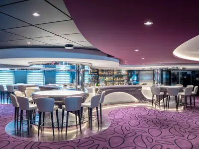 Die moderne Infinity Bar an Bord der MSC Virtuosa ist mit einer geschwungenen Theke, bequemen Barhockern, einer umfangreichen Spirituosenwand und einem auffälligen lila Teppich ausgestattet. Deckenbeleuchtung erhellt den stilvollen Raum.