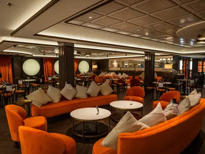 Die L'Atelier Bar & Art an Bord der MSC Virtuosa. Eine elegante Lounge mit orangefarbenen Sofas, Sesseln, Tischen und einer Bühne mit Klavier. Runde Fenster prägen den Hintergrund.