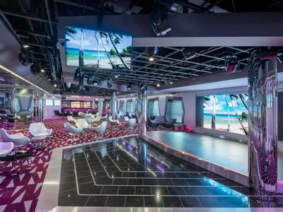 Moderne TV Studio & Bar der MSC Virtuosa mit Bühne, Lounge-Sesseln und Bar. Große Bildschirme zeigen einen tropischen Strand. Roter Teppich und glänzende Säulen.