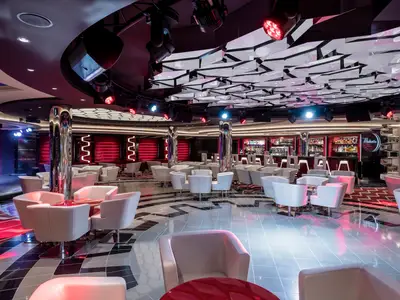 Moderne Virtuosa Bar & Lounge an Bord der MSC Virtuosa, lebhaft beleuchtet in Rot und Blau. Helle Sessel, Tische, Bühne und Bar prägen den Raum. Glänzender Boden und kunstvolle Decke schaffen elegantes Ambiente.