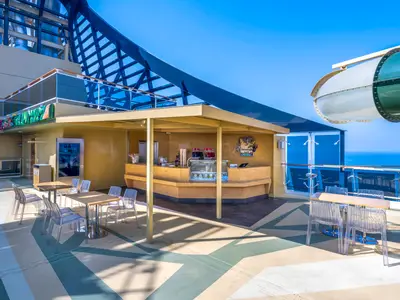 Die Savannah Bar an Bord der MSC Virtuosa bietet einen sonnigen Außenbereich mit Tischen, Stühlen und einer Bar unter einem Sonnensegel. Im Hintergrund ist das blaue Meer und eine Wasserrutsche zu sehen.
