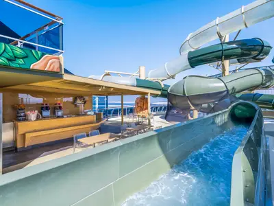 Ansicht der MSC Virtuosa mit einer Wasserrutsche, die ins Wasser führt. Links befindet sich eine Bar mit Getränkestationen und Tischen unter einem schattigen Dach, umgeben von klarem Himmel und Meer.